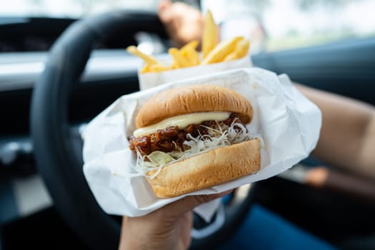 Asian lady holding hamburger and French fries to eat in car, dangerous and risk an accident.