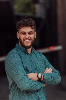 A successful young businessman in a shirt, with crossed arms, poses outdoors, confident expression on his face