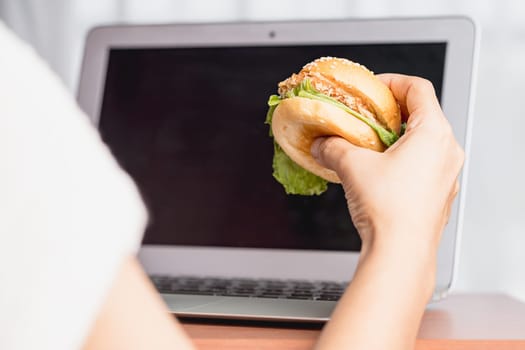 Hand holding a delicious hamburger and using laptop computer for eating while working concept.