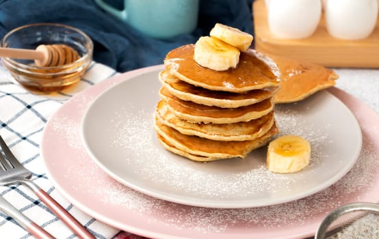 homemade banana pancakes on plate sprinkled powder and honey. Pancakes on the kitchen table against the background of eggs and cups with coffee. Pancake slide with honey topping and fresh bananas