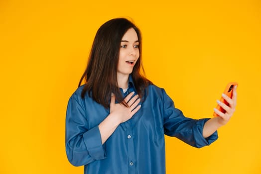 Digital Surprise: Captivating Young Woman in Stylish Shirt, Reading Astonishing News on her Phone - Vibrant Expression Isolated on Yellow Background