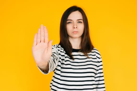 Determined Woman Making Stand: Serious Lady with Confident Expression Raising Hand in 'Stop' Gesture - Assertiveness Concept Isolated on Vibrant Yellow Background