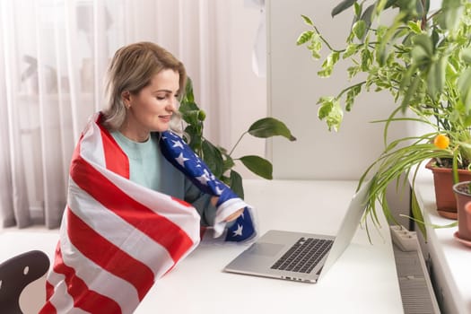 Happy woman employee sitting wrapped in USA flag, shouting for joy in office workplace, celebrating labor day or US Independence day
