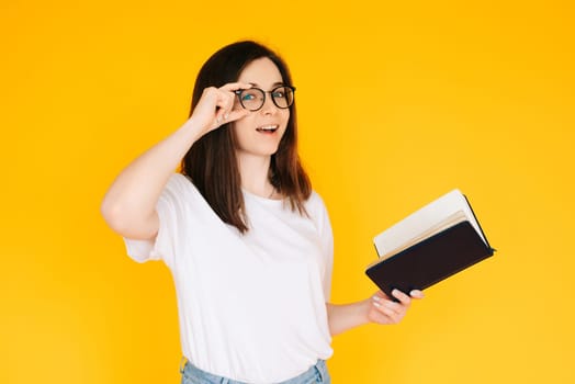 Portrait of a happy young woman wearing glasses and white T-shirt, immersed in reading a book with an open mouth, isolated on a yellow background.