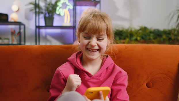 Young blonde child kid girl use mobile smartphone typing playing browsing say Wow yes found out great big surprise, good news, celebrate online game win. Female toddler at home in room sits on sofa
