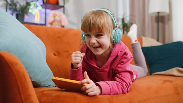 Worried funny child kid girl enthusiastically playing online racing or shooter video games on smartphone. Female teen toddler using smartphone gadget app with drive simulator at home lying on couch