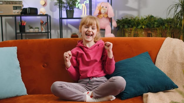 Child kid girl shouting, celebrating success, winning, goal achievement, good news, raising fists in gesture I did it, lottery luck. Female teen toddler play game at home in living room sits on sofa