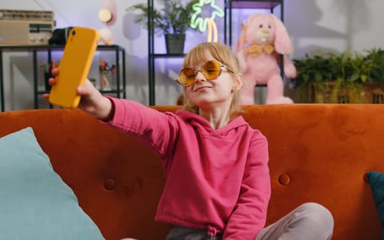 Young blonde child kid girl blogger taking selfie on smartphone, communicating video call online with social media subscribers. Female teen toddler recording video story at home in room sits on sofa