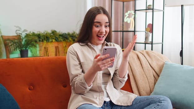 Happy young woman use mobile smartphone typing browsing say Wow yes found out great big win good news celebrate lottery jackpot doing winner gesture. Portrait of girl at home in room sitting on sofa