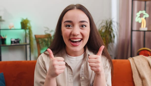 Happy young woman shouting, celebrating success, winning, goal achievement good news raising thumbs up, like gesture, lottery luck at modern home apartment. Girl in living room sitting on orange couch
