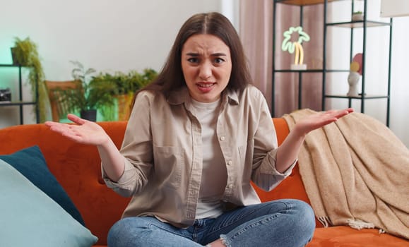 What. Why. Woman raising hands in indignant expression asking reason of failure demonstrating disbelief irritation by troubles at modern home apartment. Portrait of girl in living room sitting on sofa