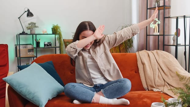 Portrait of trendy cheerful positive young woman having fun dancing and moving to rhythm, dabbing raising hands, making dub dance gesture sitting on sofa. Girl at modern home living room apartment