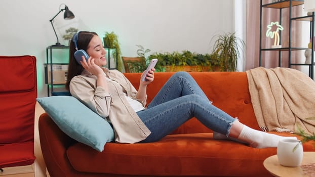 Portrait of happy overjoyed woman in wireless headphones relaxing lying on couch at home apartment choosing listening favorite energetic disco dancing music. People weekend daytime leisure activities
