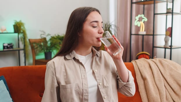 Thirsty young woman holding glass of natural aqua make sips drinking still water preventing dehydration sits at home living room. Girl with good life habits, healthy slimming, weight loss concept