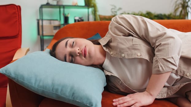 Tired young woman lying down in bed taking a rest at home living room apartment. Carefree girl napping, falling asleep on comfortable sofa with pillows. Closed her eyes enjoy daytime nap alone on sofa