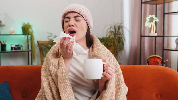 Young ill woman suffering from cold or allergy sitting on sofa in living room at home drinking hot tea. Sick brunette girl wear hat wrapped in plaid blows sneezes wipes snot into napkin. Quarantine