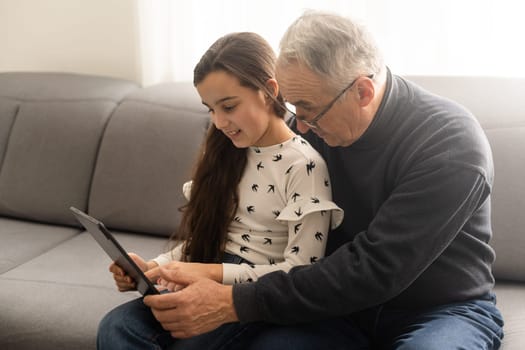 Cute little girl and her handsome grandpa are smiling while sitting on couch at home. Girl is using a tablet