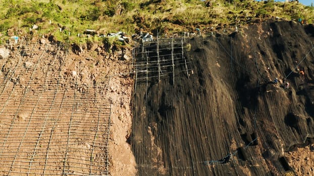 Workers strengthen the slope of the mountain with metal mesh preventing rockfall and landslide on the road, above view. workers constructing anti-landslide concrete wall prevent protect against rock slides. safety concept