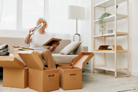 elderly woman sits on a sofa at home with boxes. collecting things with memories albums with photos and photo frames moving to a new place cleaning things and a happy smile. Lifestyle retirement. High quality photo