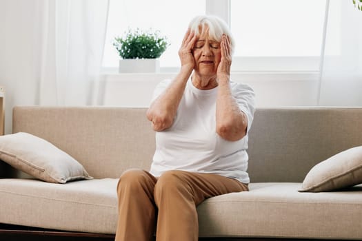 Elderly woman severe pain in the head sitting on the couch, health problems in old age, poor quality of life. Grandmother with gray hair holding her head, migraine and high blood pressure. High quality photo