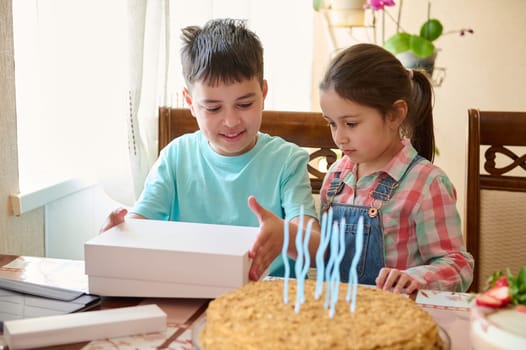Authentic portrait of Caucasian adorable boy opening birthday present, sitting at table with a birthday cake. Little girl and teen boy celebrating birthday event. People. Lifestyle. Childhood