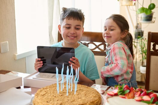 Adorable teen boy smiles rejoicing at a digital tablet for his birthday, sitting at table with delicious homemade cake, celebrating his birthday. People. Lifestyle. Technology. Life events. Childhood