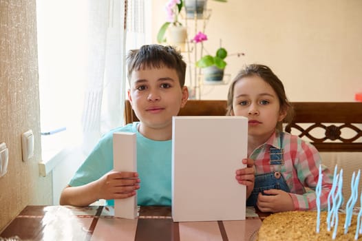 Authentic portrait of a teen boy and his younger sister holding white mockup box , sitting at table and celebrating happy life event. White gift boxes with copy space for advertising text
