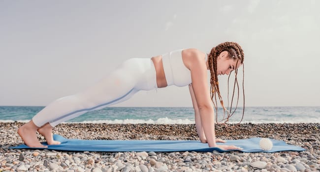 Woman yoga sea. Well looking middle aged woman with braids dreadlocks in white leggings and tops doing stretching pilates on yoga mat near sea. Female fitness yoga routine concept. Healthy lifestyle