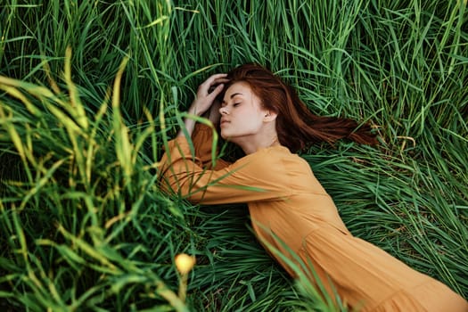 a close horizontal photo of a pleasant woman in a long orange dress resting lying in the tall grass with her eyes closed in sunny weather with her arms outstretched. High quality photo