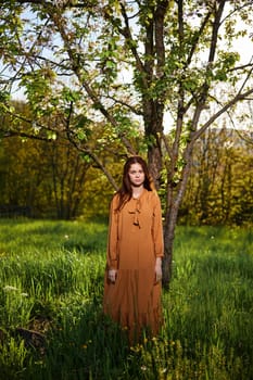 a sweet, attractive woman with long red hair stands in the countryside near a flowering tree in a long orange dress and looks at the camera with a slight smile on her face. High quality photo