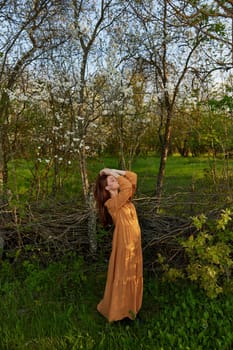 an elegant, sophisticated woman poses relaxed standing near a wicker fence at the dacha in a long orange dress touching her head with her hands. High quality photo