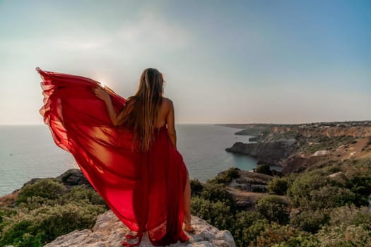 Woman sunset sea red dress, back view a happy beautiful sensual woman in a red long dress posing on a rock high above the sea on sunset