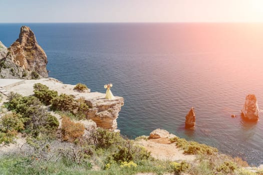 Woman yellow dress sea. Side view Young beautiful sensual woman in yellow long dress posing on a rock high above the sea at sunset. Girl in nature against the blue sky.