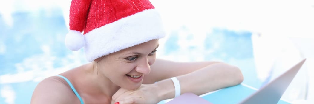 Woman in red santa claus hat looking at laptop on sunbed. Removed merry christmas and happy new year greetings concept