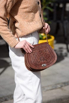 woman in white pants and a beige sweater poses outside with a small leather handbag.