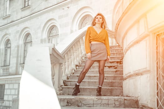 A middle-aged woman looks like a good blonde with curly beautiful hair and makeup on the background of the building. She is wearing a yellow sweater