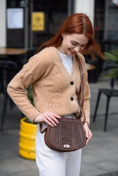 a red-haired girl in white pants and a beige sweater poses outside with a small leather handbag.