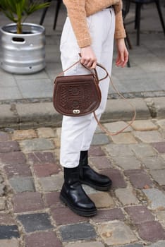 woman in white pants and a beige sweater poses outside with a small leather handbag.