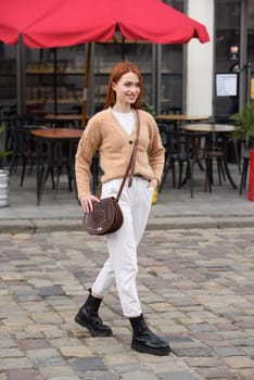 a red-haired girl in white pants and a beige sweater poses outside with a small leather handbag.