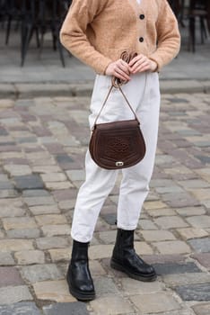 woman in white pants and a beige sweater poses outside with a small leather handbag.