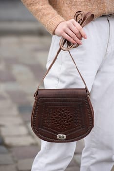 woman in white pants and a beige sweater poses outside with a small leather handbag.