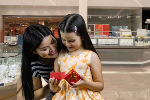 Little girl opens a red gift box from her mother. Birthday celebration concept. Woman and her daughter in the mall.