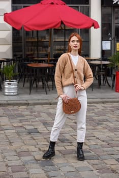 a red-haired girl in white pants and a beige sweater poses outside with a small leather handbag.