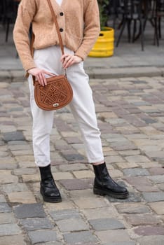 woman in white pants and a beige sweater poses outside with a small leather handbag.