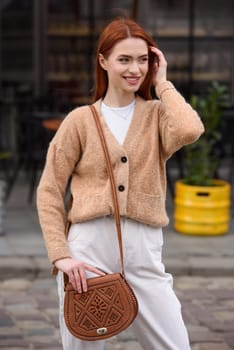 a red-haired girl in white pants and a beige sweater poses outside with a small leather handbag.