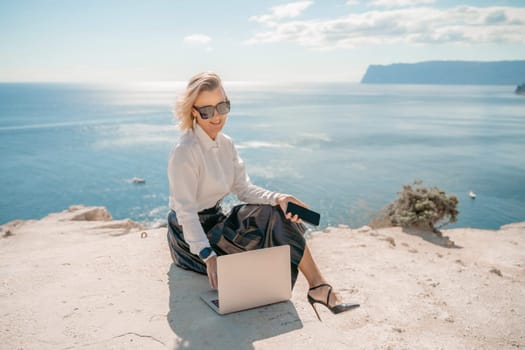 Freelance women sea working on a computer. Pretty middle aged woman with computer and phone outdoors with beautiful sea view. The concept of remote work