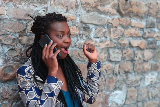 confident African young adult woman holding cell phone calling , receives good news, smiling, happy, outdoors in the village over a wall.