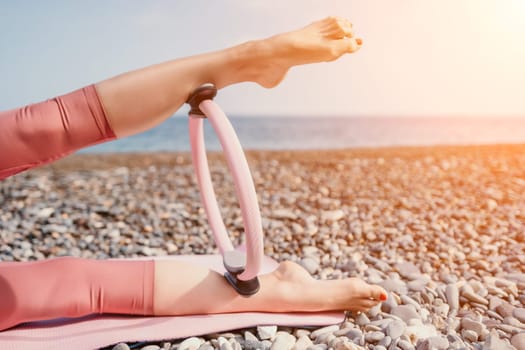 Middle aged well looking woman with black hair doing Pilates with the ring on the yoga mat near the sea on the pebble beach. Female fitness yoga concept. Healthy lifestyle, harmony and meditation.