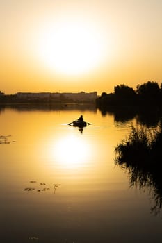 A young man is fishing on a lake from an inflatable small boat at a very beautiful orange sunset