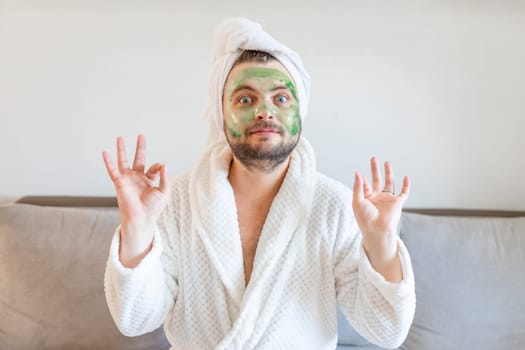Funny man enjoys beauty treatments and luxury relaxation at home spa. A man with a towel on his head sits on a sofa in a white bathrobe and with a cosmetic mask on his face